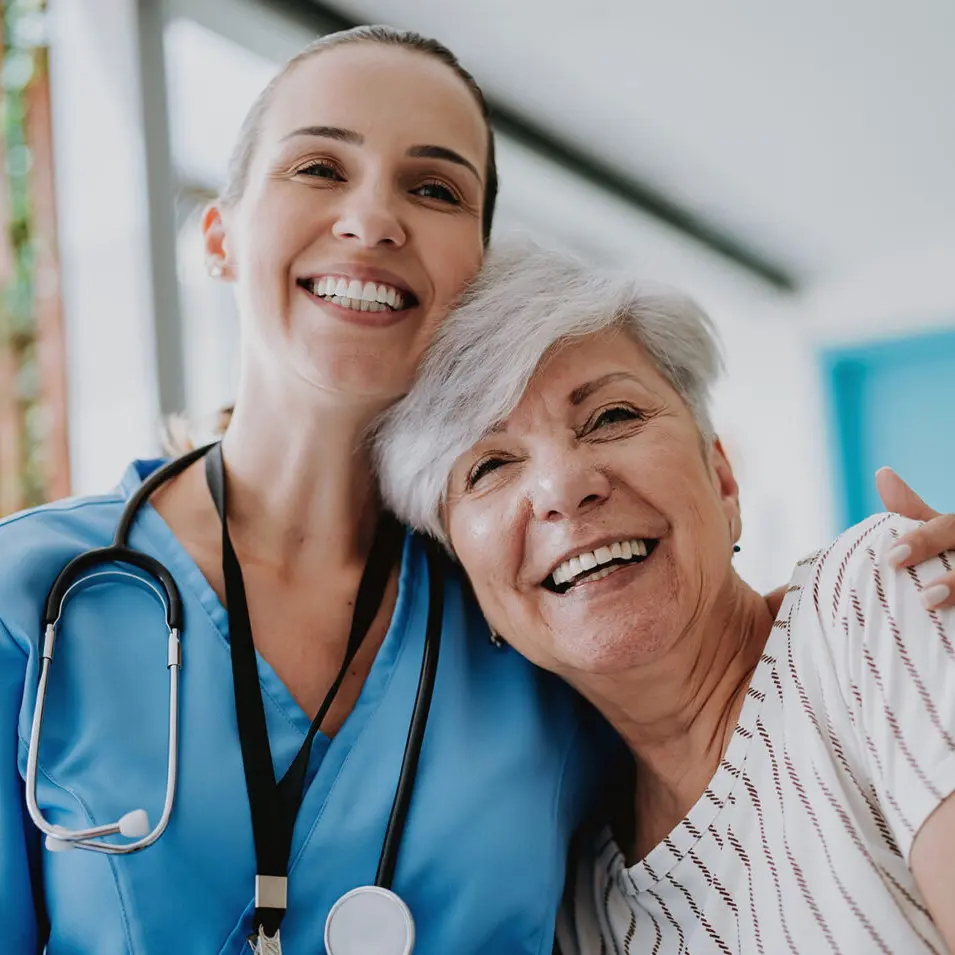 Smiling nurse with a stethoscope embracing an older patient, both looking happy in a bright healthcare setting.